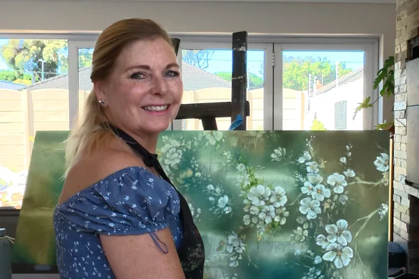 Linda Wolf in her studio, smiling, seated beside a canvas of white blossoms on green.