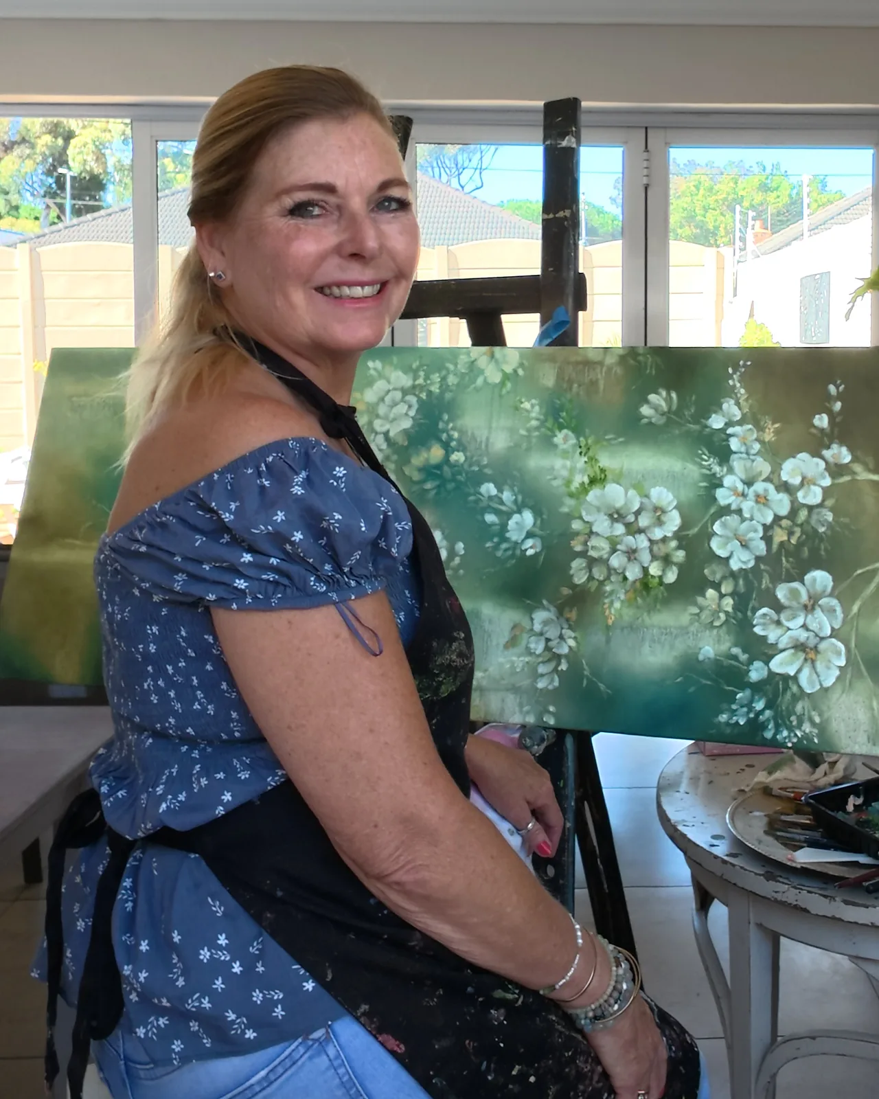 Linda seated beside a canvas of white blossoms on green, smiling.
